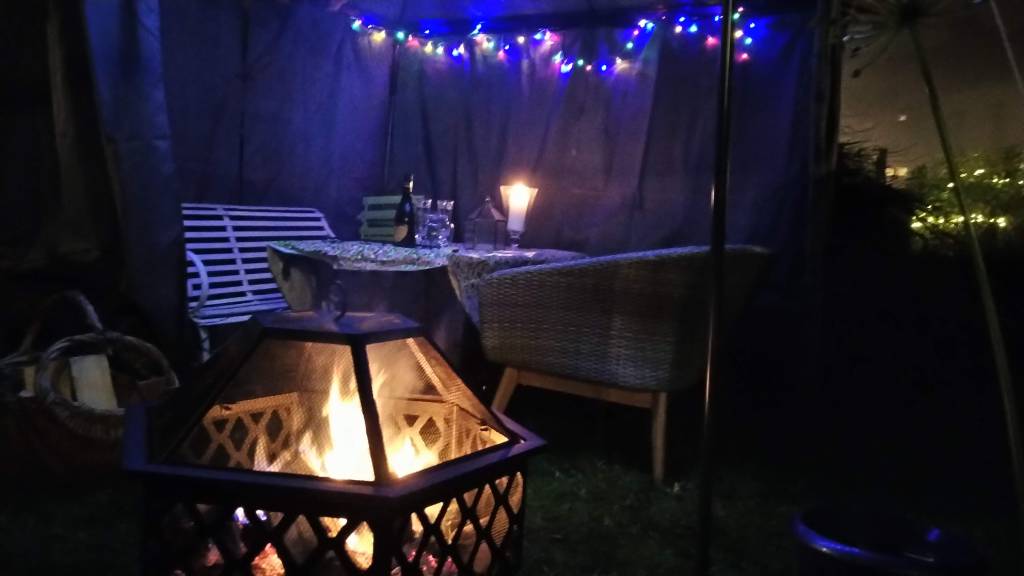 Fire bowl with fire, gazebo with coloured fairy lights, and a bottle of prosecco on a table between garden seating