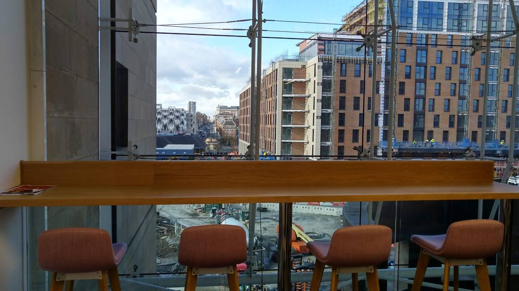 Four chairs look out of a plat glass window. Outside a building site, and city in the distance