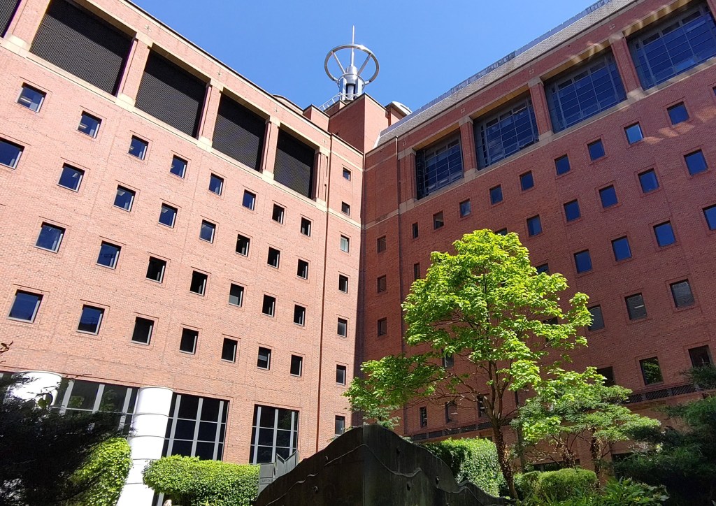 Garden with shrubs and trees in front of 7-storey red brick office building