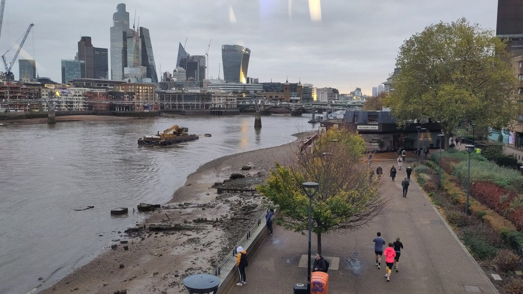 View across wide footpath next to muddle Thames waterside. In the distance are city of London skyscrapers