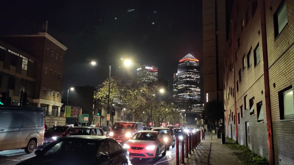 Night-time vew through queues of traffic towards Canary Wharf. Two skyscraper towers with many lights on, one of which has a pyramid roof
