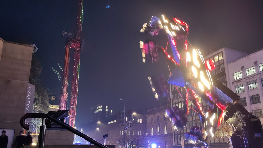 Night-time scene, steps to art gallery on left, on the right, a fairground ride is brightly lit whirling people around high in the air