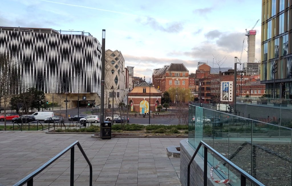 city centre buildings from left - a modern multi-storey car park clad in metal fins that are twisted to make a gothic arch pattern, a small octagonal brick building in the middle of a traffic roundabout, the end of a row of mid-20th-century brick offices, the concrete lift core of a skyscraper under construction, the edge of a glass-walled building. In the foreground a pair of railings for steps which are out of shot