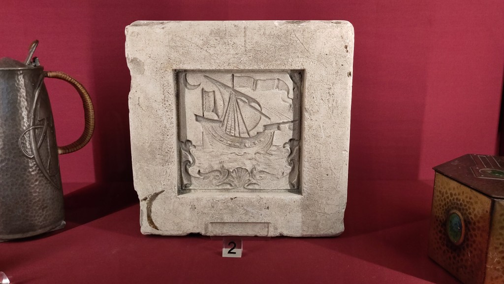 Mould for a ceramic tile with indented square featuring a sailing boat design. The tile is in a museum display case between a metal jug and metal tin