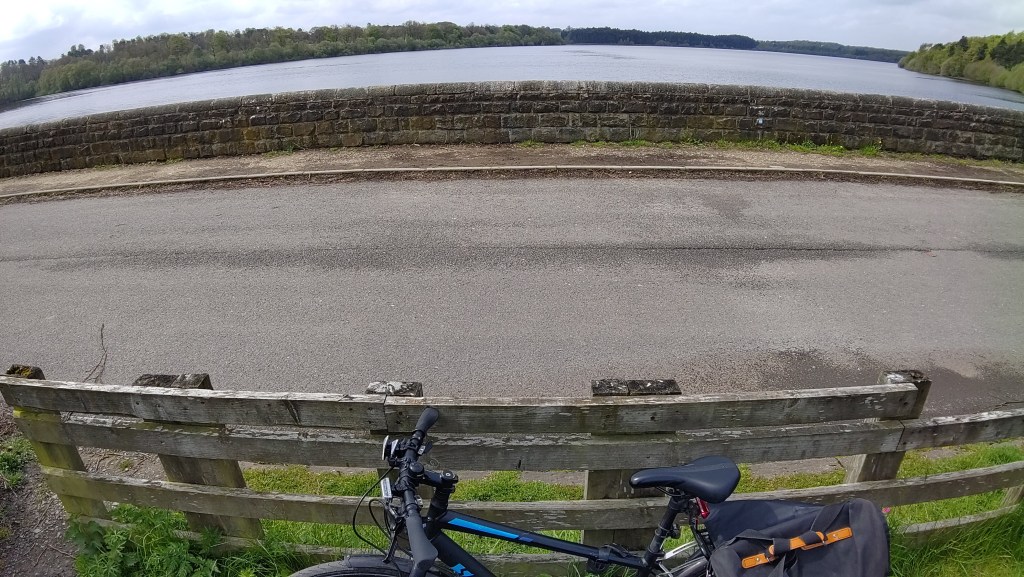From bottom (foreground) to top (far distance):The top of a hybrid bicycle with pannier The bike is leaning on a wooden fence A single track road A stone wall A large expanse of water Trees line the banks on the far side of the water