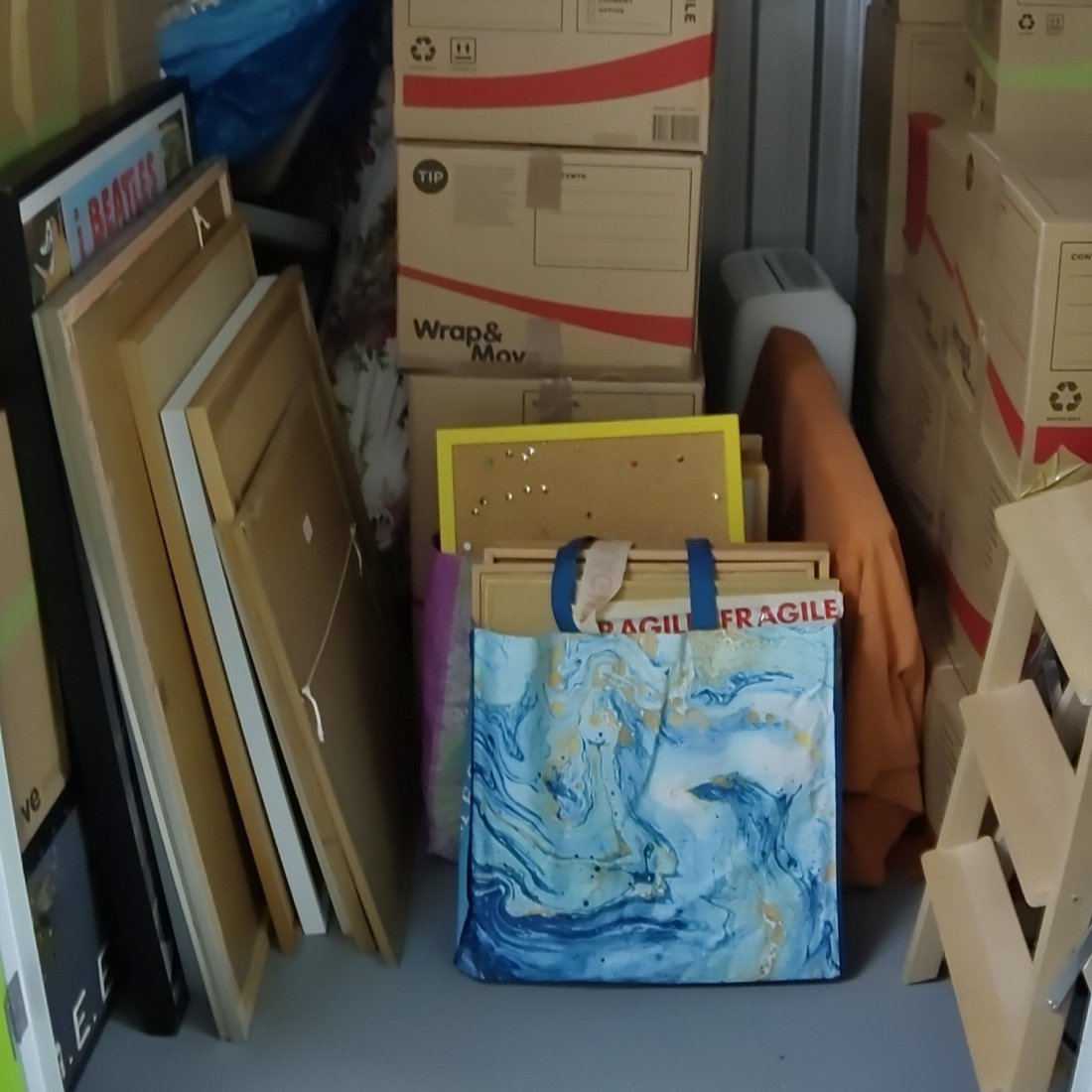 Piles of household objects including pictures leaning in a stack facing the wall, piles of cardboard boxes, a pinboard, a small wooden step-ladder, a dehumidifier. In the foreground, a "bag for life" with a marbled effect contains smaller pictures. The back of one of the pictures has "FRAGILE" tape on it