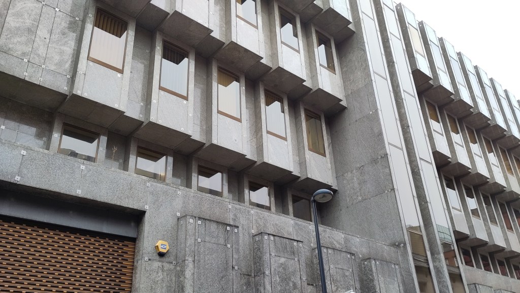 A grey marble clad facade with each of four floors cantilvered out beyond the floor below. Regularly spaced portrait shaped windows stand proud of the facade adding further to the blocky overall impression
