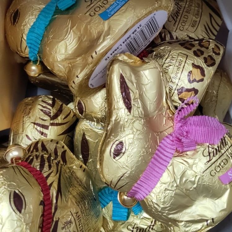 Close-up of a jumble of Lindt chocolate bunnies. They are covered in gold foil with rabbit eyes, ears, and whiskers printed in brown. Each bunny wears a coloured ribbon as a collar with a little bell on it