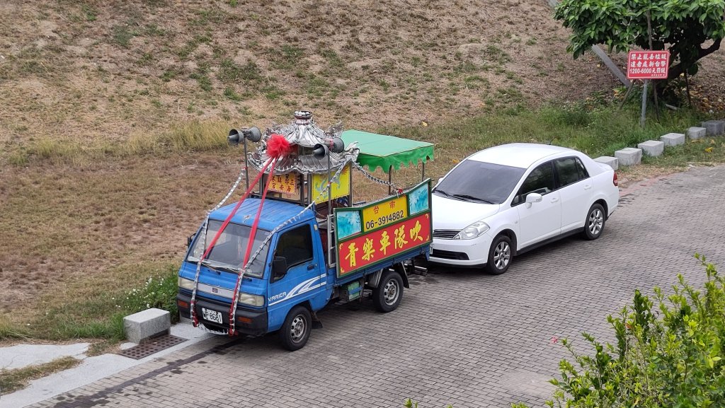 Looking down at a road with two parked vehicles. One is an ordinary white car. The other is a small blue cabbed truck with a traditional Chinese structure on top, with loudhailers either side pointing front and back. Behind the vehicles, a dry, scrubby grass bank slopes upwards