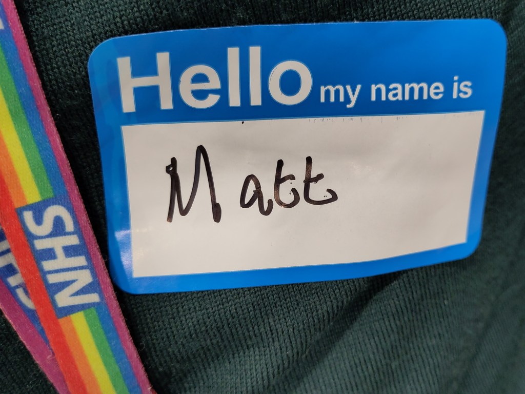 A name sticker that says "Hello my name is" with "Matt" written in a box in black marker pen. Next to it, a section if rainbow-coloured lanyard with the NHS lozenge. Both on a dark green knitted background