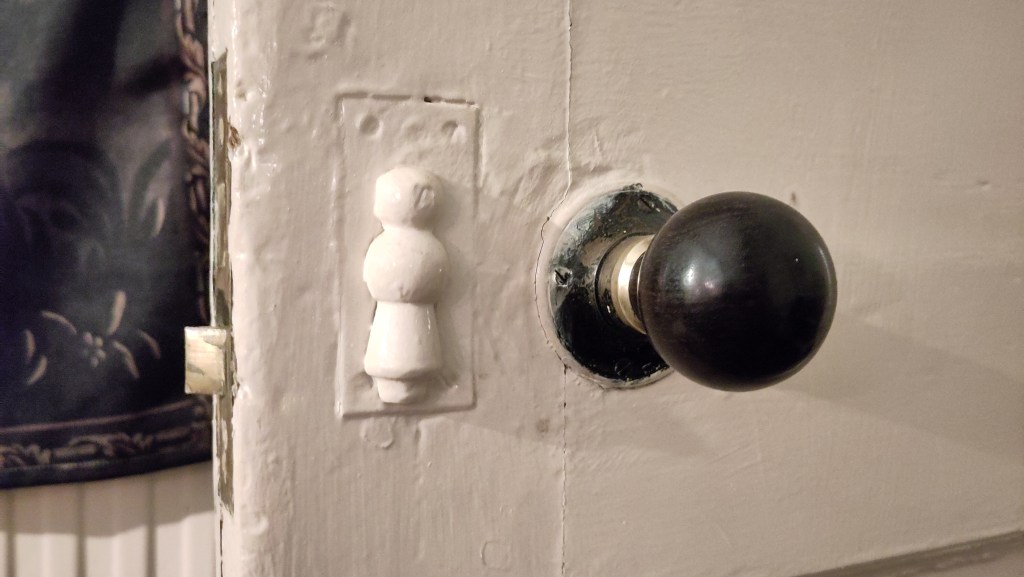 Close-up of a round wooden doorknob on a thick white gloss painted wooden door. To the left of it a lock cover is painted up. Behind the door, the corrugations of a radiator are visible