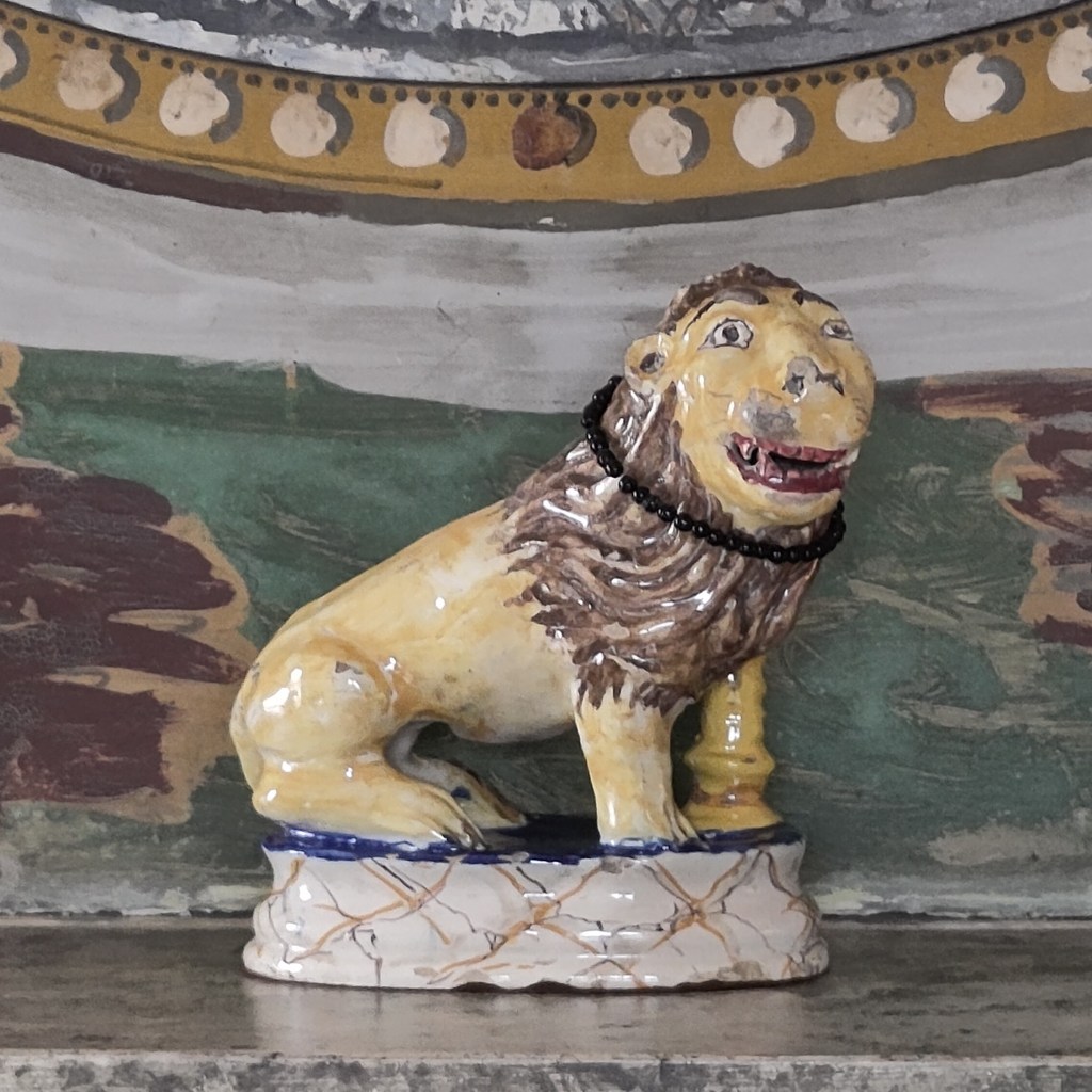 A ceramic lion on a marble mantle shelf. The lion is naively made with an almost human face, and glazed in with yellow body and face and brown mane. The glaze is chipped in places. Around its neck is a circle of small back beads. Behind the lion, the wall is roughly but richly painted in earthy green, brown, yellow, grey and white.
