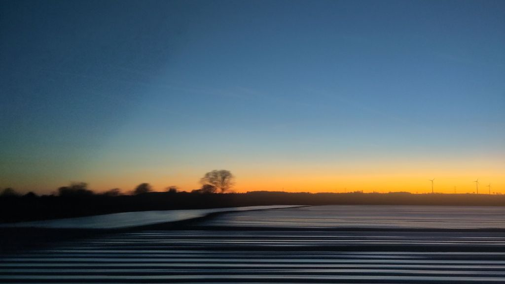 Sunset seen from a moving train. In the foreground horizontal marks across a ploughed field, at the horizon a row of about 5 trees, slightly out of focus, one larger than the rest. The sky is a gradient from orange at the bottom to dusty deep blue at the top