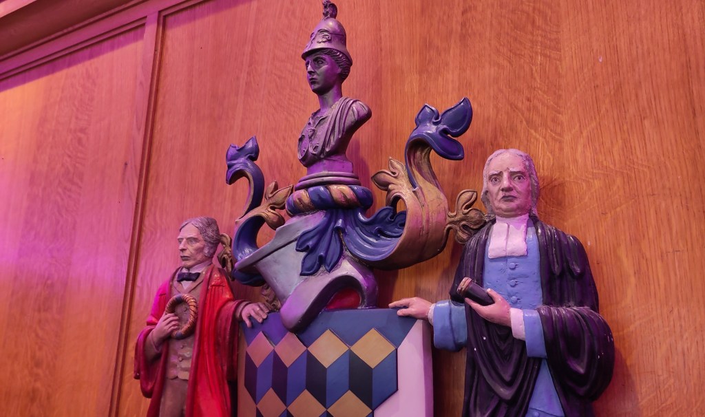 A painted relief carved crest mounted on a wood panelled wall. The top half of a shield shows a repeating patterns of black, blue and gold cubes. Supporters either side of the shield are men wearing historic clothing and gowns. Above the shield, a helmet with a classical head and shoulders bust on top