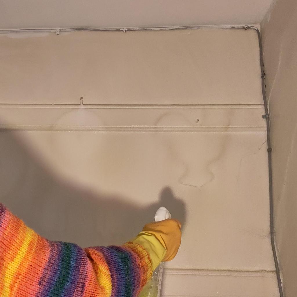 An arm wearing a stripy jumper is holding a spray bottle of cleaning fluid. The spray is pointed at a wall with off white paint covered in years of grime. In the grime are outlines or objects that used to be hung on the wall, including the shape of a chicken